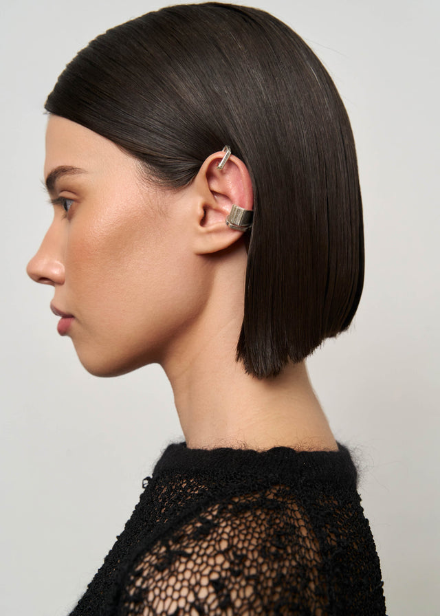 Profile portrait: two ribbed silver ear cuffs on the ear, dark bob, black knit, grey studio backdrop — STRUGA