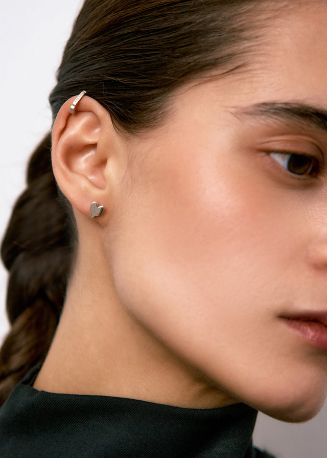 Silver heart stud in the lobe with a slim ear cuff above it, ear seen in profile — STRUGA