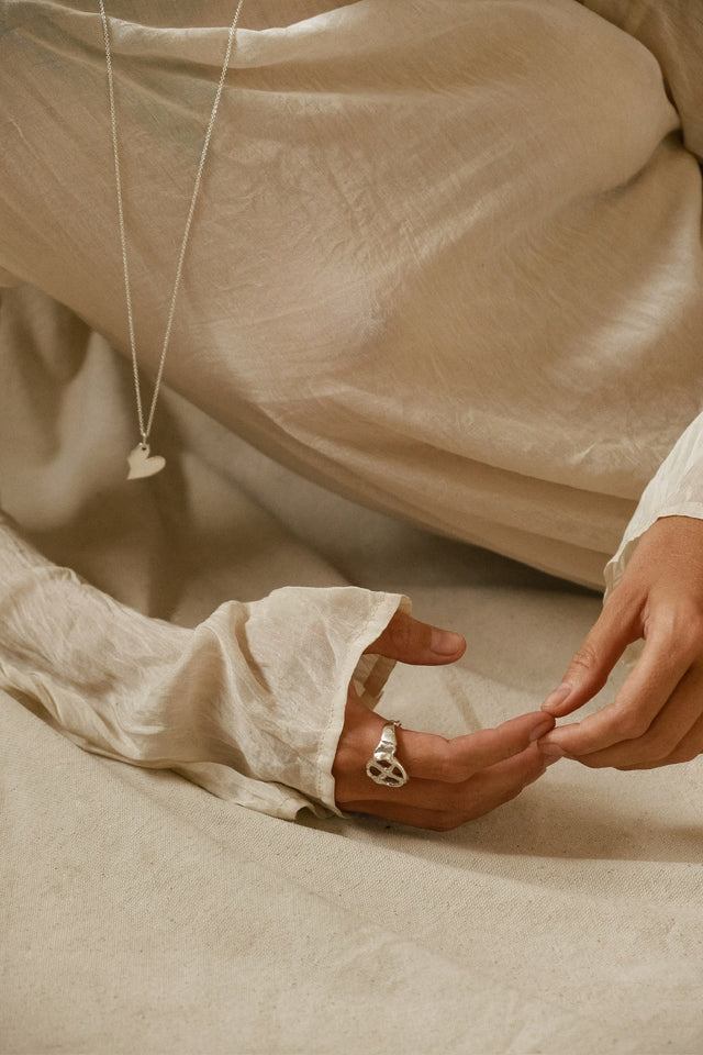Hand in a silver open-cross ring, two fingertips touching over cream draped fabric, heart pendant on a chain — STRUGA