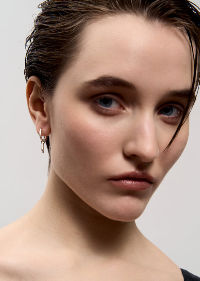 Close portrait: silver huggie hoop earring with a small cylinder drop in the earlobe, soft pale grey backdrop — STRUGA
