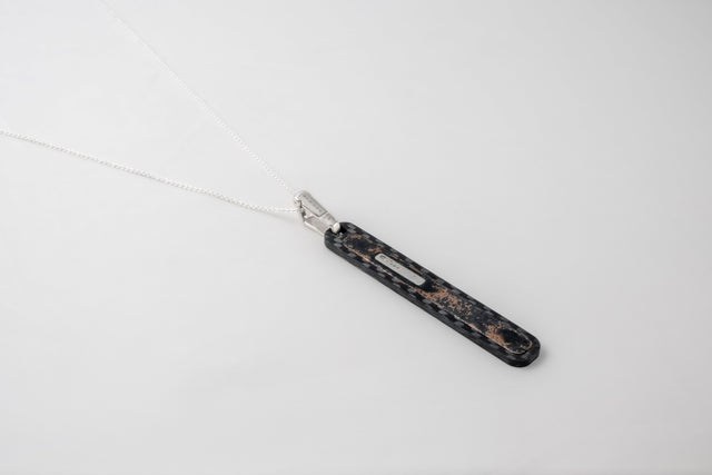 Long dark carbon pendant with copper flecks on a fine silver chain, laid on a pale ground.