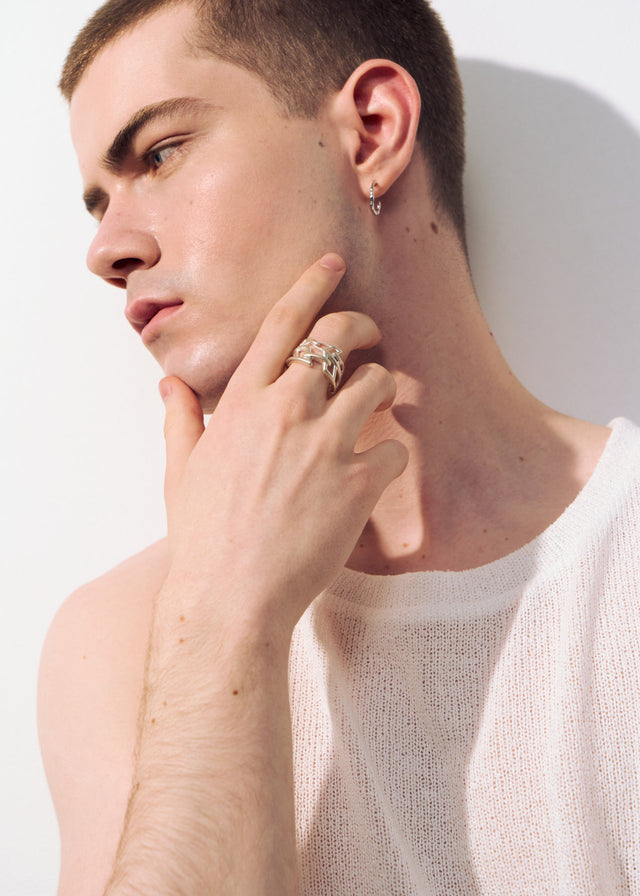 Wide silver chain-link ring on the index finger, hand lifted to the jaw, small silver ear hoop, pale studio wall — STRUGA
