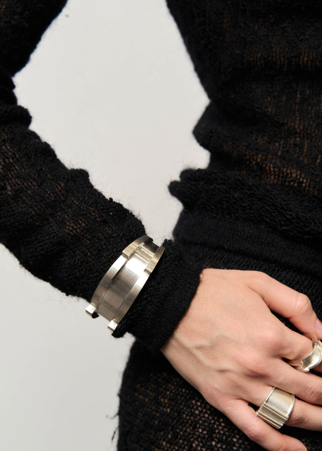 Wide brushed silver cuff on a wrist against a black knit sleeve, rings on the hand beside it — STRUGA