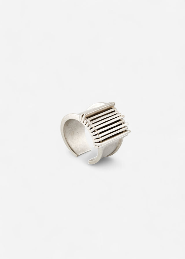 Open cuff band in cool white metal, its outer face a grille of parallel raised bars, plain pale studio backdrop — STRUGA