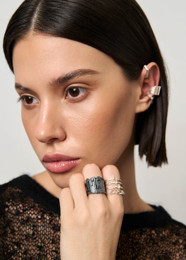 Blackened silver Brutalism V.3 ring on a hand at the chin, silver ear cuffs above, light ground — STRUGA