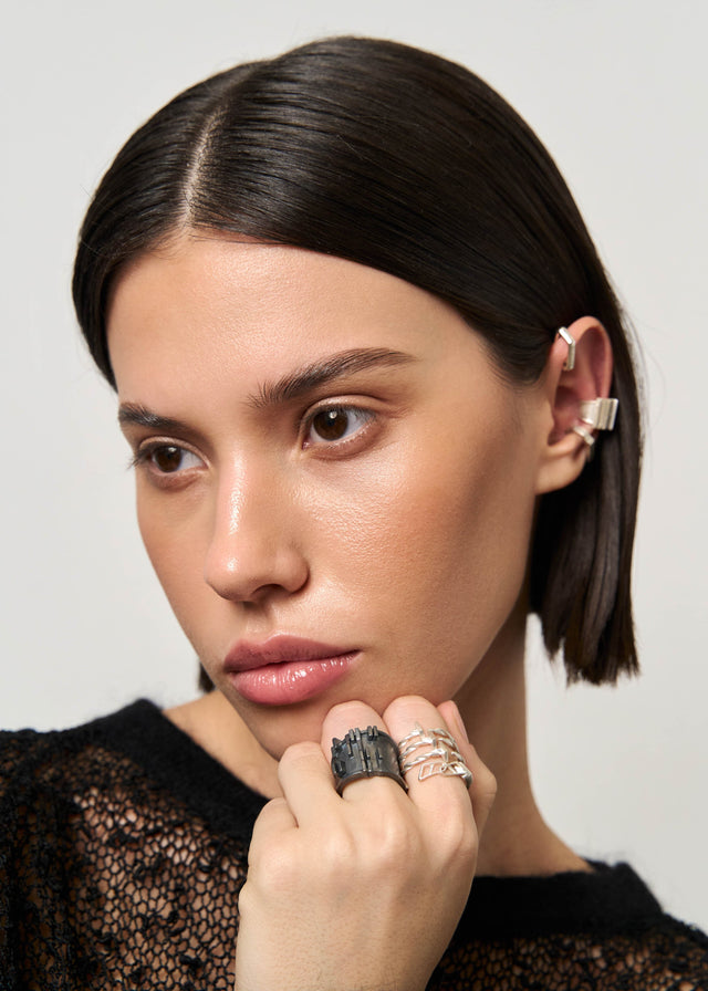 Blackened and bright silver rings on one hand, silver ear cuffs above, black open-knit top — STRUGA