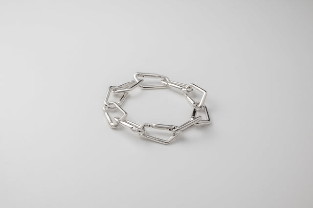 Polished silver bracelet of angular elongated links, coiled flat on a pale grey studio backdrop — STRUGA
