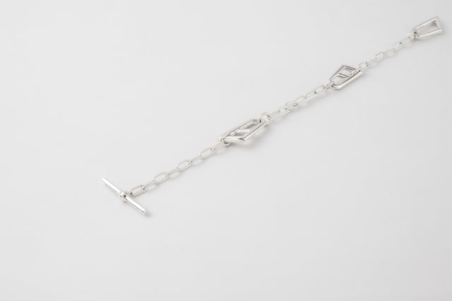 Polished silver chain bracelet laid out flat on a pale grey backdrop, flat geometric links and T-bar clasp — STRUGA