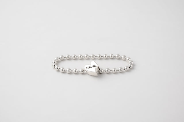 Polished silver ball-bead bracelet with an engraved heart bead in the chain, laid flat on a pale grey backdrop — STRUGA