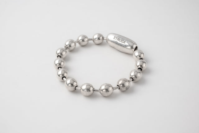 Chunky ball-link silver bracelet with an engraved oval capsule clasp, lit on a pale grey backdrop — STRUGA