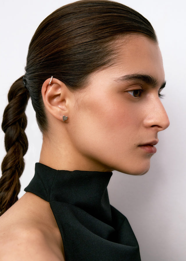 Profile view: silver-toned heart stud in the lobe and slim bar ear cuff on the upper ear, pale studio backdrop — STRUGA