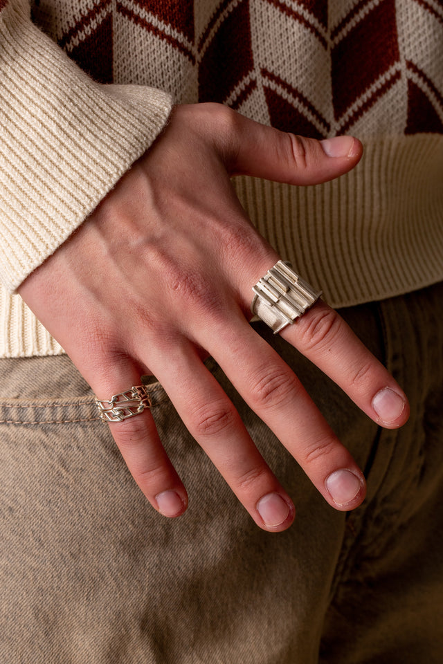 Hand from above on a tan trouser pocket, wearing a wide ridged silver band and an openwork silver link ring — STRUGA