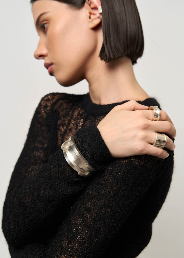 Model in black open-knit sweater, two wide silver rings and a broad silver cuff, pale grey studio backdrop — STRUGA