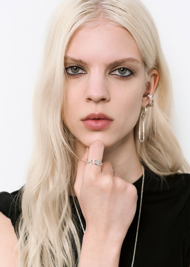 Blonde model in black top wears an open silver chain-link ring on her index finger, hand at her chin, white studio — STRUGA